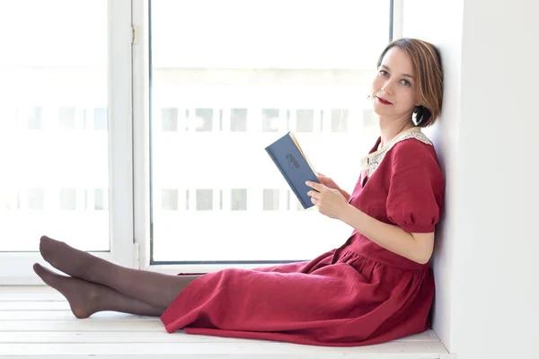 Portrét nádherné okouzlující mladé dívky ve skromných červených šatech, které čtou knížku u okna. Koncept domácího pohodlí čtení knih a lásky k klasice. — Stock fotografie