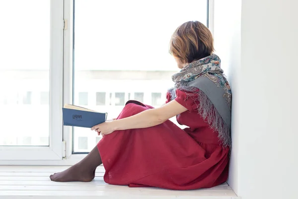 Vista laterale di una ragazza affascinante con un abito lungo e modesto e una sciarpa sta leggendo un libro mentre è seduto sul davanzale della finestra. Concetto di gioventù e romanticismo . — Foto Stock