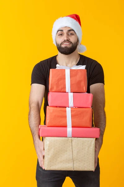 Niezadowolony młody człowiek z brodą w kapeluszu Świętego Mikołaja posiada pięć pudełek upominkowych stwarzających na żółtym tle. Koncepcja prezentów i pozdrowienia dla świąt Bożego Narodzenia i nowego roku. — Zdjęcie stockowe