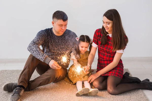 Концепція святкування, сім'ї та свят - щасливі батьки та маленька донька з блискавками сидять на килимі на сірому фоні — стокове фото