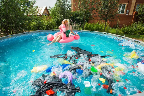 Probleem van afval, plastic recycling, vervuiling en milieu concept-domme vrouw zwemt en plezier hebben in een vervuild zwembad. Flessen en plastic zakken zweven in de buurt van haar — Stockfoto
