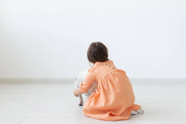 Liten flicka med mörkt hår leker med sin älskade hund när man sitter på golvet i en persika klänning mot bakgrund av vita väggen. Konceptet att ta hand om barn och djur. Reklamplats. — Stockfoto