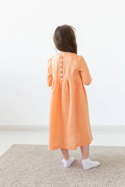 Rückansicht eines kleinen Mädchens, das in einem langen pfirsichfarbenen Kleid mit Knöpfen auf dem Rücken und weißem Hintergrund posiert. das Konzept der einzigartigen Kinderkleider. Werbefläche. — Stockfoto