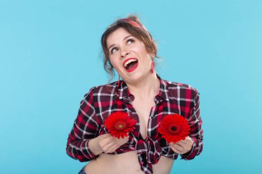 Ekose gömlek li pozitif genç kadın portresi mavi arka plan karşı poz meme yakınında farklı kalpleri ile iki kırmızı çiçek tutan. Her türlü vücut güzelliği kavramı, Vücut pozitifliği