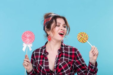 Elinde renkli lolipop tutan ve mavi arka planda poz birini yalayan retro giysiler içinde Büyüleyici genç kadın. Tatlılar ve tatiller için aşk kavramı.