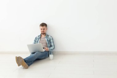 Boş bir odada yerde oturan yeni konut arayışı içinde Internette sörf Casual giysiler olumlu genç adam. Internet ve dizüstü bilgisayar kullanarak bir daire bulma kavramı. Boşaltmak