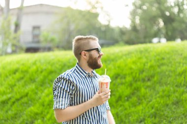 Gözlük ve şık sakallı pozitif genç hipster adam bir parkın karşısında dışarıda dururken bir saman ile milkshake içiyor. Hafif tatlı içecekler sevenler kavramı.