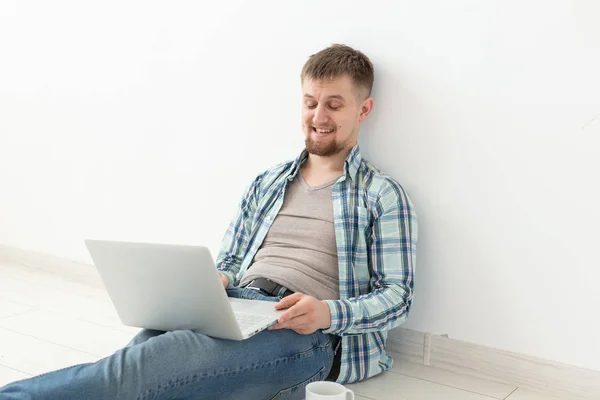 Pozytywny młody człowiek w casual ubrania surfowanie po Internecie w poszukiwaniu nowych mieszkań siedzi na podłodze w pustym pomieszczeniu. Koncepcja znalezienia mieszkania za pomocą Internetu i laptopa. Miejsce — Zdjęcie stockowe