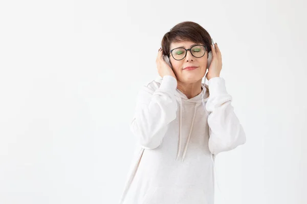 Linda mujer de mediana edad positiva en suéter y gafas está escuchando música con auriculares de alambre de pie sobre un fondo blanco con espacio para copiar. Concepto de pasatiempos y suscripciones a la radio favorita — Foto de Stock