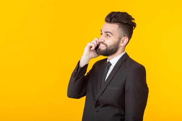 Cute young man with a beard in formal clothes talking on the phone posing on a yellow background with copy space. Concept of an informal meeting and business deal.