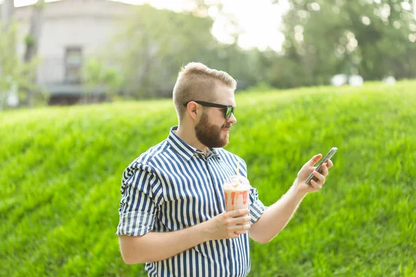 El hipster masculino joven y positivo escucha su radio en línea favorita con auriculares para teléfonos inteligentes e Internet inalámbrico de alta velocidad en el parque en un día soleado y cálido de verano . — Foto de Stock