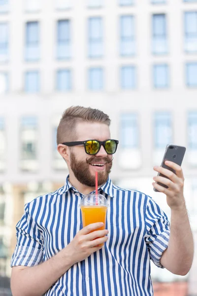 Вид сбоку на молодого стильного хипстера в очках, с усами и соком в руках, болтающего в социальных сетях с помощью смартфона и беспроводного интернета в теплый летний день . — стоковое фото