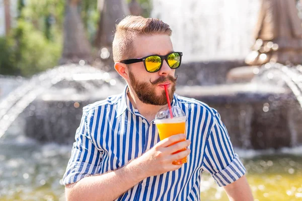 Lindo hipster joven y elegante con bigote y barba y gafas bebiendo jugo con una pajita mientras está sentado en un parque de la ciudad sobre el fondo de una fuente. Concepto vacaciones de verano . — Foto de Stock