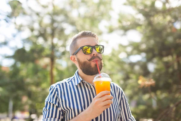 Portret van een positieve vrolijke jongeman met een glas sap met een rietje tijdens het wandelen in het Park op een warme zonnige zomerdag. Het concept van rust na studie en werk in het weekend. — Stockfoto