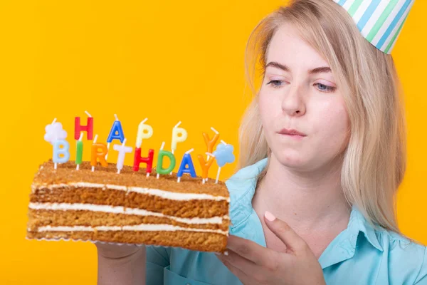 Ritratto ravvicinato di una divertente ragazza positiva con un cappuccio di carta che tiene una torta di congratulazioni tra le mani su uno sfondo giallo. Concetto e divertimento e festa. Spazio pubblicitario . — Foto Stock