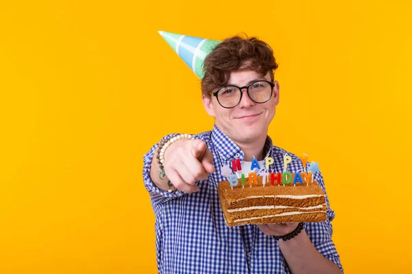 Divertente ragazzo positivo tiene in mano una torta fatta in casa con l'iscrizione buon compleanno in posa su uno sfondo giallo. Concetto di vacanze e anniversari . — Foto Stock