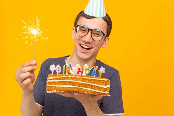 Divertente ragazzo positivo in occhiali tiene tra le mani una torta fatta in casa con l'iscrizione buon compleanno in posa su uno sfondo giallo. Concetto di vacanze e anniversari . — Foto Stock