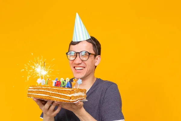 Loco joven alegre asiático con gafas sosteniendo una vela ardiente en sus manos y un pastel casero de felicitación sobre un fondo amarillo con espacio para copiar. Celebración de cumpleaños y aniversario —  Fotos de Stock