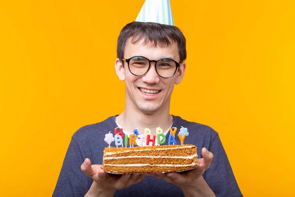 Divertente ragazzo positivo in occhiali tiene tra le mani una torta fatta in casa con l'iscrizione buon compleanno in posa su uno sfondo giallo. Concetto di vacanze e anniversari . — Foto Stock