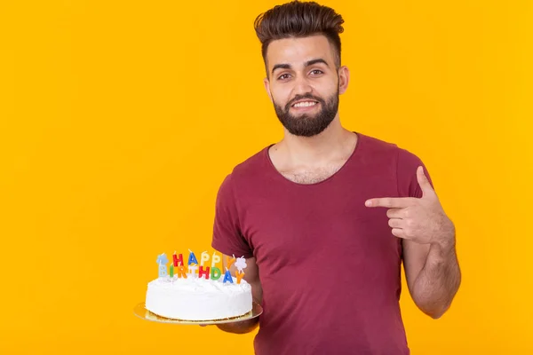 Homem hipster bonito positivo na camiseta de Borgonha que mantém o bolo congratulatório com a inscrição feliz aniversário posando em um fundo amarelo. Conceito de parabéns e aniversários . — Fotografia de Stock