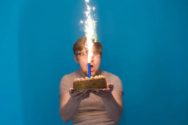 Безумный веселый парень в очках здоровается с бумажной шляпой, держа торт с горящим фейерверком в руках. День рождения — стоковое фото