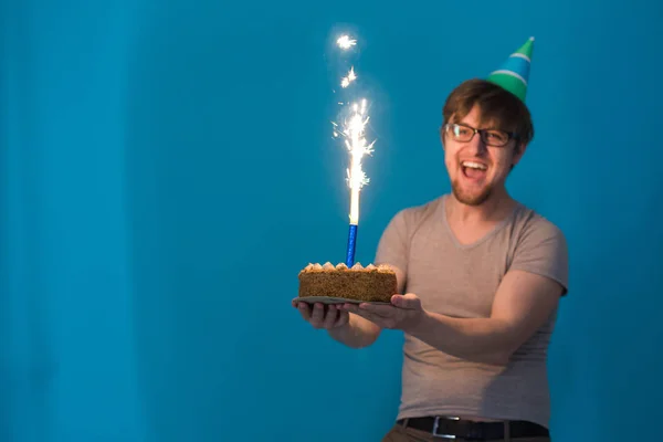 Божевільний веселий хлопець студент в окулярах вітається з паперовим капелюхом, тримаючи торт з палаючим феєрверком в руках. Концепція святкового дня народження — стокове фото
