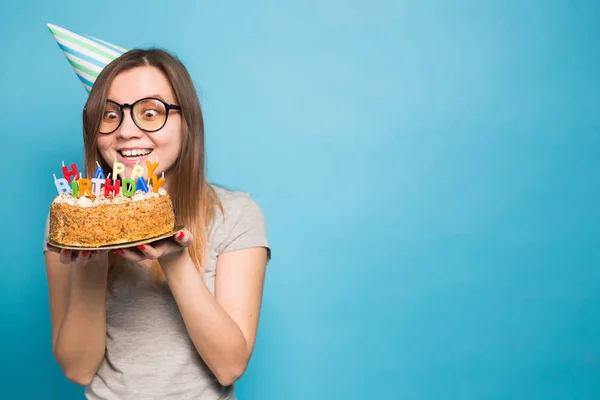 파란색 배경에 큰 생일 케이크를 들고 종이 모자와 안경에 미친 재미 소녀. 장난과 인사의 개념. — 스톡 사진