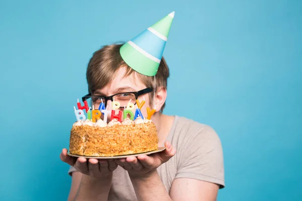 メガネと紙のお祝いの帽子でクレイジー面白い若者は、コピースペースと青い背景に立ってケーキ幸せな誕生日を保持 — ストック写真