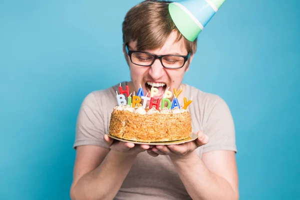 Divertido macho en papel sombrero de felicitación tratando de morder un pastel con un feliz cumpleaños velas de pie sobre un fondo azul . —  Fotos de Stock