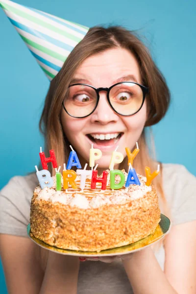 Loca chica divertida en un sombrero de papel y vasos sosteniendo un gran pastel de cumpleaños en el fondo azul. Concepto de broma y saludos . — Foto de Stock