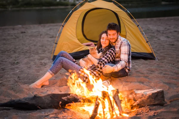 People, tourism and nature concept - Man and woman with their cat sit near a fire