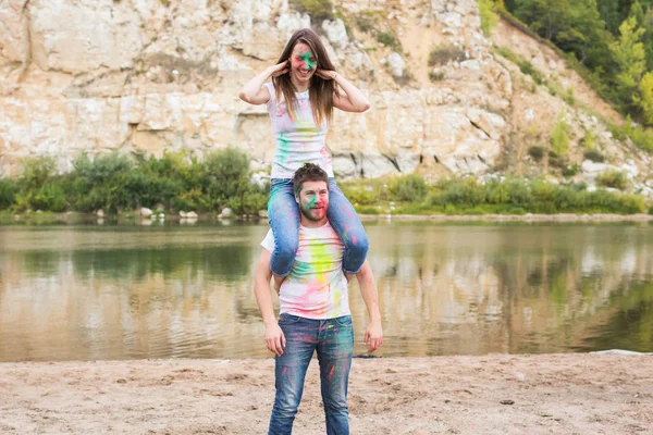 Festival holi, turismo de verão e conceito de natureza - jovem mulher atraente sentado piggyback em seu namorado na natureza — Fotografia de Stock