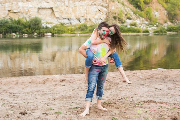 Festival holi, turismo de verão e conceito de natureza - jovem mulher atraente sentado piggyback em seu namorado na natureza — Fotografia de Stock