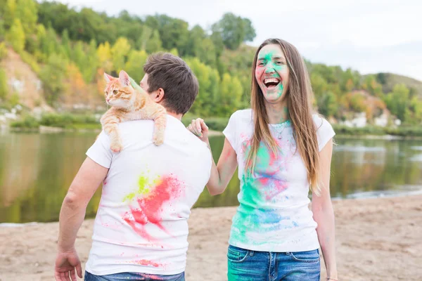 Festival holi, vacanze, turismo, pet e natura concetto - Ritratto di donna e uomo con gatto coperto polvere multicolore — Foto Stock