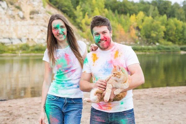 Festival holi, feriados, turismo, animal de estimação e conceito de natureza - Retrato de mulher e homem com gato coberto de poeira multicolorida — Fotografia de Stock