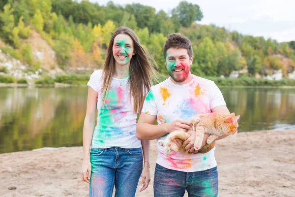 Festival holi, holidays, tourism and nature concept - Couple dressed in white shirts holding cat and covered with colorful dust — Stock Photo, Image