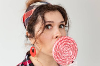 Şişkin gözleri ile Komik güzel genç kadın beyaz bir arka plan üzerinde poz renkli lolipop ile ağzını kapatır. Aşk konsepti ve yüksek kalorili tatlılar.