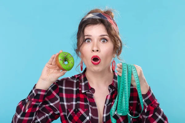 Close-up shocked surprised young beautiful woman looking at measuring tape and holding a green donut in hands posing on a blue background. Concept of diet and rejection of harmful high-calorie foods.