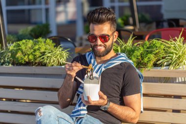 Güneşli bir yaz gününde bir park bankında otururken bir öğle yemeği kutusundan Çin erişte yeme genç yakışıklı hipster adam. Sağlıklı ve besleyici bir atıştırmalık kavramı.