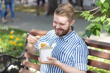 Güneşli bir yaz gününde bir park bankında otururken bir öğle yemeği kutusundan Çin erişte yiyen genç yakışıklı hipster adam Yan görünümü. Sağlıklı ve besleyici bir atıştırmalık kavramı.
