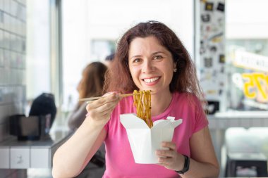 Bir kafede Çin eriştesi yiyen ve pencereden dışarı bakan genç ve neşeli bir kızın portresi. Sağlıklı Asya mutfağı kavramı.