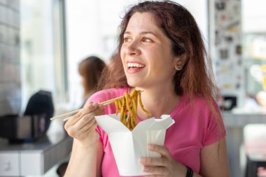 Bir kafede Çin eriştesi yiyen ve pencereden dışarı bakan genç ve neşeli bir kızın portresi. Sağlıklı Asya mutfağı kavramı.