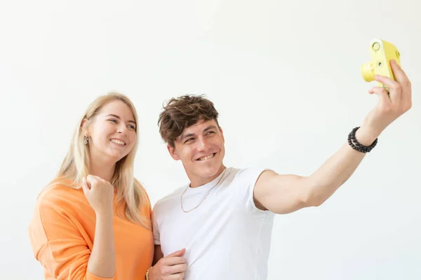 Engraçado louco jovem casal loira e um cara hipster tomando uma selfie em uma câmera de filme amarelo vintage posando em um fundo branco. Conceito de hobby de fotografia . — Fotografia de Stock