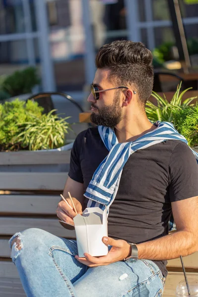 Positieve jonge stijlvolle Arabische kerel die Chinese noedels in een park eet tijdens een pauze op het werk. Het concept van rust en gezonde snack — Stockfoto