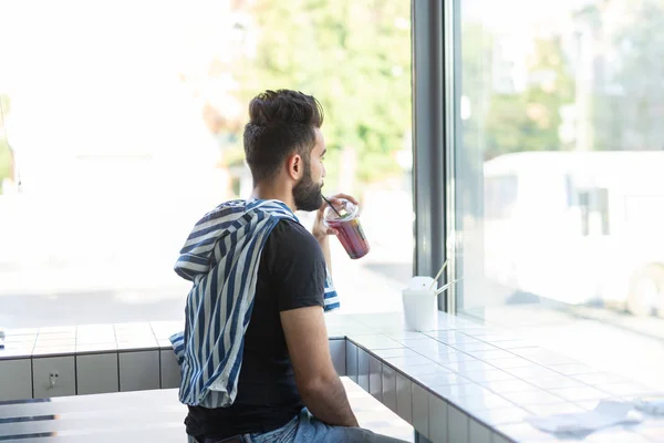 Handsome young hipster guy drinking cocktail in a cafe and looking into the window.
