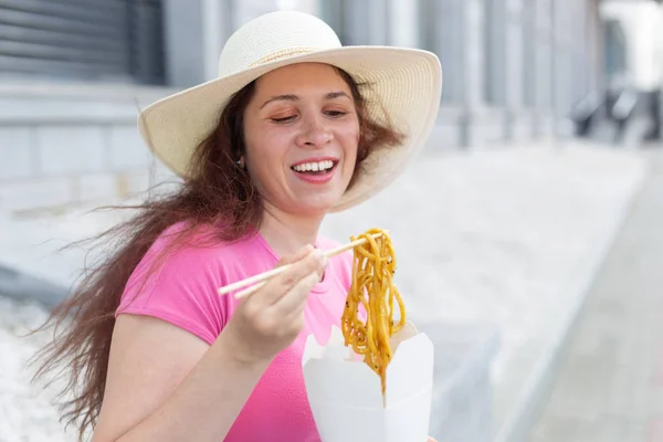 Joven chica positiva en un sombrero come fideos chinos con palillos de madera para el almuerzo para llevar. Concepto de comida saludable alta en calorías . — Foto de Stock