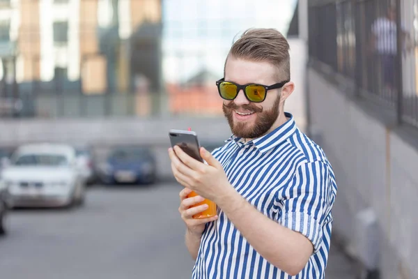 Widok z boku młodego stylowego Hipster człowiek z okularami i wąsy i sok w ręce rozmowy w sieciach społecznościowych za pomocą smartfona i bezprzewodowego Internetu w ciepłym letnim dniu. — Zdjęcie stockowe