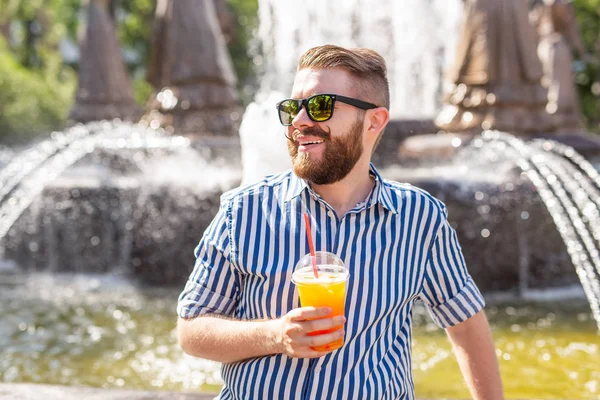 口ひげとあごひげを持つかわいいスタイリッシュな若い男性ヒップスターと噴水の背景に都市公園に座っている間、わらでジュースを飲んでメガネを着用。夏休みのコンセプト. — ストック写真