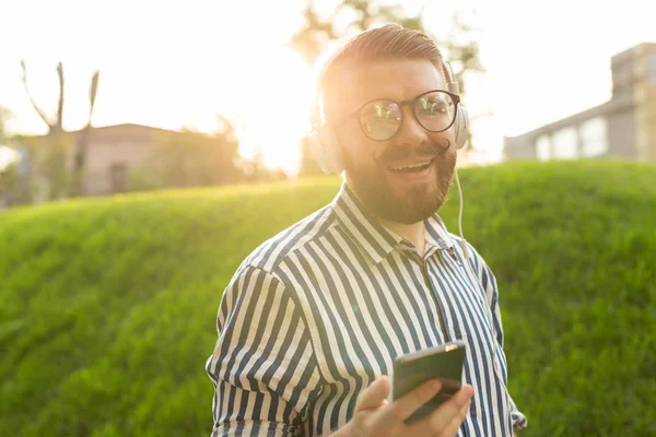 Retrato de un joven positivo con bigote y barba escuchando un audiolibro mientras paseaba por el parque. Concepto de aprendizaje del ocio . — Foto de Stock