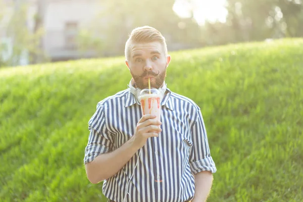 Positive young hipster guy in glasses and stylish beard is drinking a milkshake with a straw while standing outside against a park. The concept of lovers of light sweet drinks.